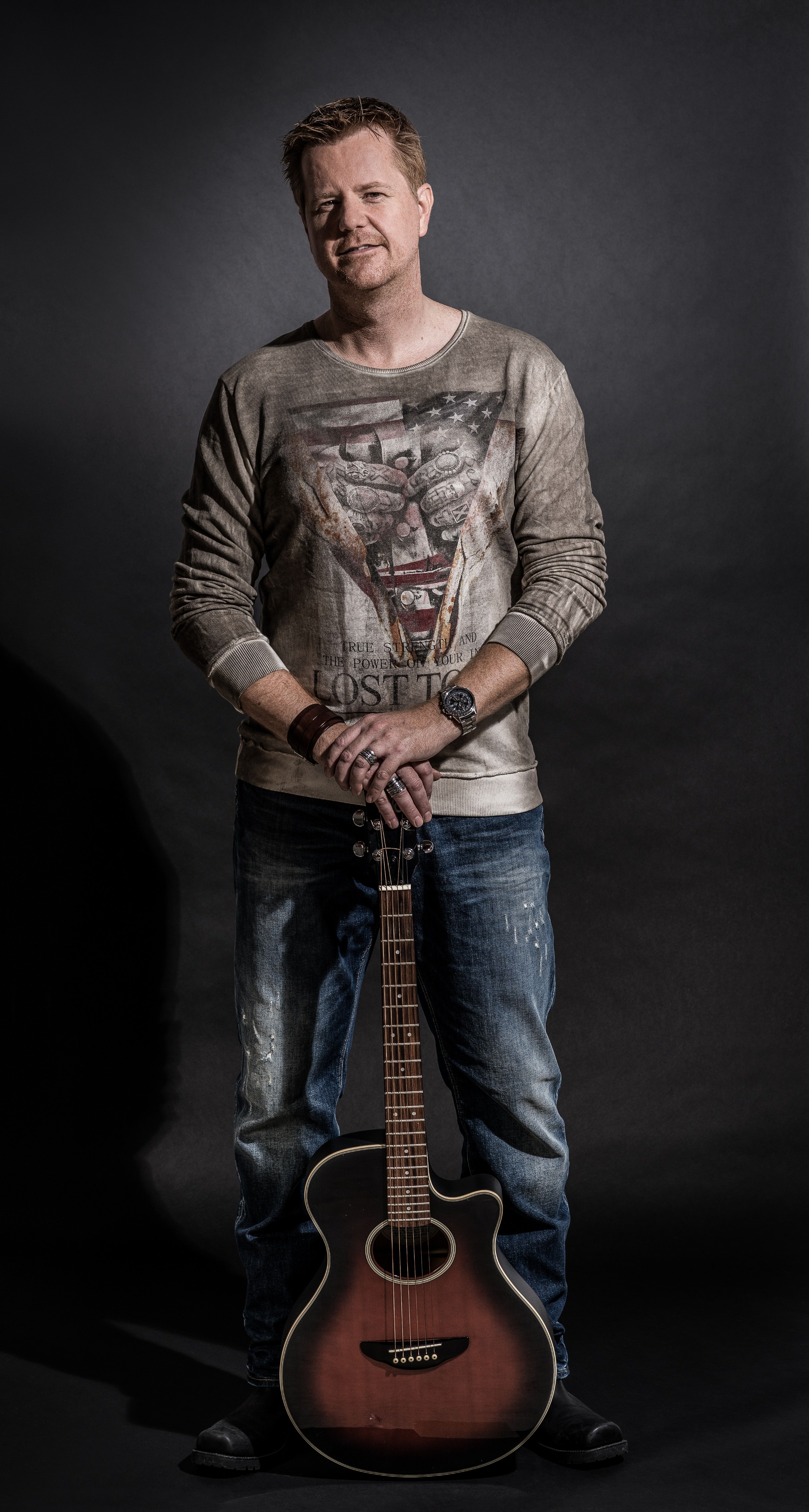 Ben Valdern steht in Jeans und Sweatshirt vor einem dunklen Hintergrund. Vor ihm steht eine Gitarre auf dem Boden, die er mit beiden Händen festhält.